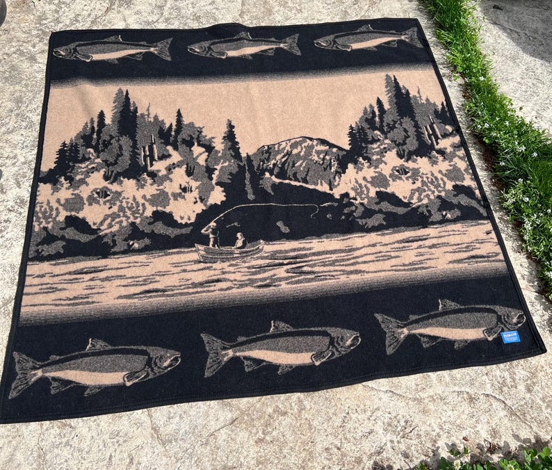 Puede incluir: Una manta tejida rectangular con un esquema de color negro y bronceado. El dise&ntilde;o muestra un paisaje central de un lago, un peque&ntilde;o bote con dos figuras y monta&ntilde;as. Los bordes superior e inferior presentan un patr&oacute;n repetitivo de peces.