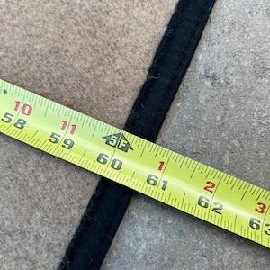 Op de afbeelding: Een beige stoffen oppervlak met een dikke, zwart gestikte rand. Een geel meetlint ligt over de stof en geeft een afmeting van ongeveer 155 cm aan bij de rand van de zwarte boord.