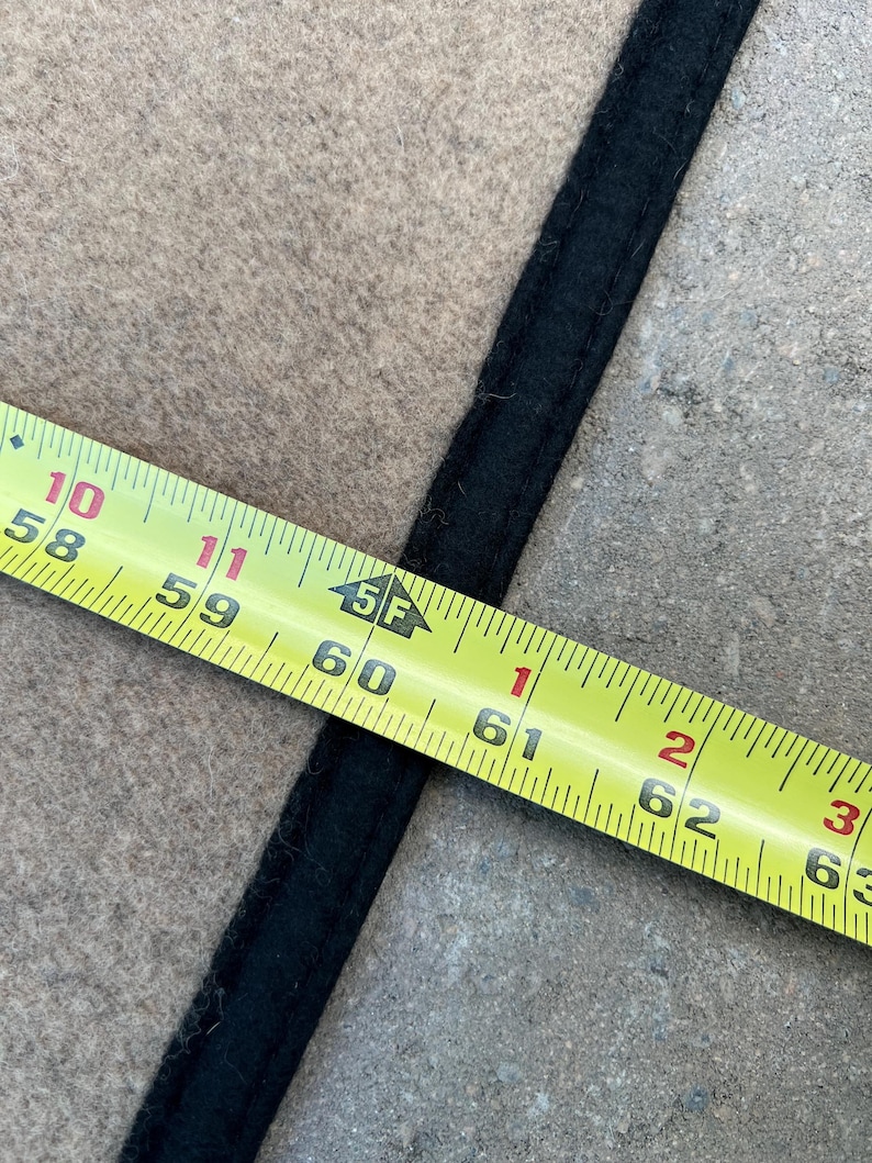 Op de afbeelding: Een beige stoffen oppervlak met een dikke, zwart gestikte rand. Een geel meetlint ligt over de stof en geeft een afmeting van ongeveer 155 cm aan bij de rand van de zwarte boord.