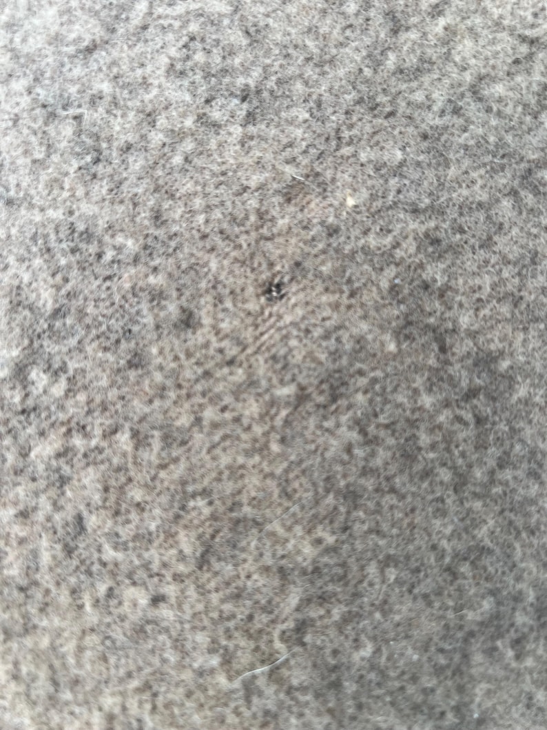 Op de afbeelding: Een close-up van een grijs viltwollen oppervlak met een dichte, matte textuur. Het materiaal vertoont een gem&ecirc;leerd uiterlijk met variaties in licht- en donkergrijze vezels, inclusief een klein, donker, cirkelvormig gaatje in de stof.
