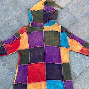 K&ouml;nnte beinhalten: Ein lang&auml;rmeliger Kapuzenpullover mit einem Patchwork-Design aus quadratischen Stoffbahnen in leuchtenden Farben wie Rot, Orange, Gelb, Violett, Blau und Olivgr&uuml;n. Das Kleidungsst&uuml;ck hat eine spitze Kapuze und sichtbare, offene N&auml;hte.