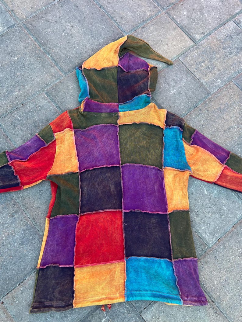 K&ouml;nnte beinhalten: Ein lang&auml;rmeliger Kapuzenpullover mit einem Patchwork-Design aus quadratischen Stoffbahnen in leuchtenden Farben wie Rot, Orange, Gelb, Violett, Blau und Olivgr&uuml;n. Das Kleidungsst&uuml;ck hat eine spitze Kapuze und sichtbare, offene N&auml;hte.