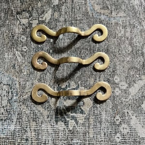 May include: Three identical brass drawer pulls arranged vertically. Each pull features a central arched handle section that curves outward, terminating in two symmetrical, outward-facing scroll ends. The metal has a smooth, polished finish.