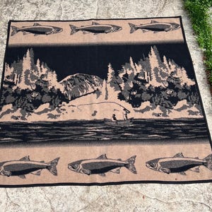 Puede incluir: Una manta tejida rectangular con un dise&ntilde;o en negro y beige. El panel central muestra un paisaje con una persona pescando desde una barca en un lago, rodeada de &aacute;rboles y monta&ntilde;as. Los bordes superior e inferior muestran filas de peces.