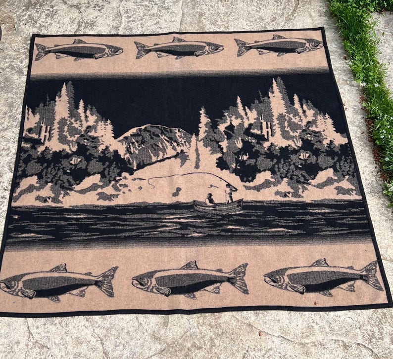 Puede incluir: Una manta tejida rectangular con un dise&ntilde;o en negro y beige. El panel central muestra un paisaje con una persona pescando desde una barca en un lago, rodeada de &aacute;rboles y monta&ntilde;as. Los bordes superior e inferior muestran filas de peces.