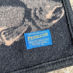 Puede incluir: Una manta de lana negra con un gr&aacute;fico grande de un pez de color beige. Una etiqueta rectangular de tela azul est&aacute; cosida en la superficie, mostrando el texto: Warranted to be a Pendleton, Pendleton Woolen Mills, Portland, Oregon, Since 1863.