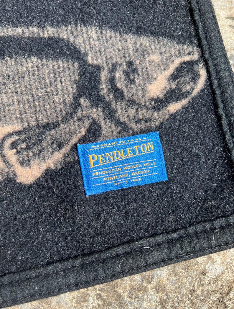 Puede incluir: Una manta de lana negra con un gr&aacute;fico grande de un pez de color beige. Una etiqueta rectangular de tela azul est&aacute; cosida en la superficie, mostrando el texto: Warranted to be a Pendleton, Pendleton Woolen Mills, Portland, Oregon, Since 1863.