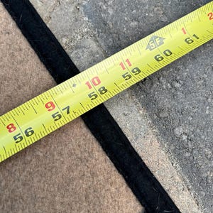 Op de afbeelding: Een beige textiel met een zwarte rand ligt onder een geel meetlint. Het lint toont markeringen van 142 tot 155 cm, wat de lengte van de rand van het beige materiaal aangeeft terwijl het uitgelijnd is met het meetinstrument in de afbeelding.