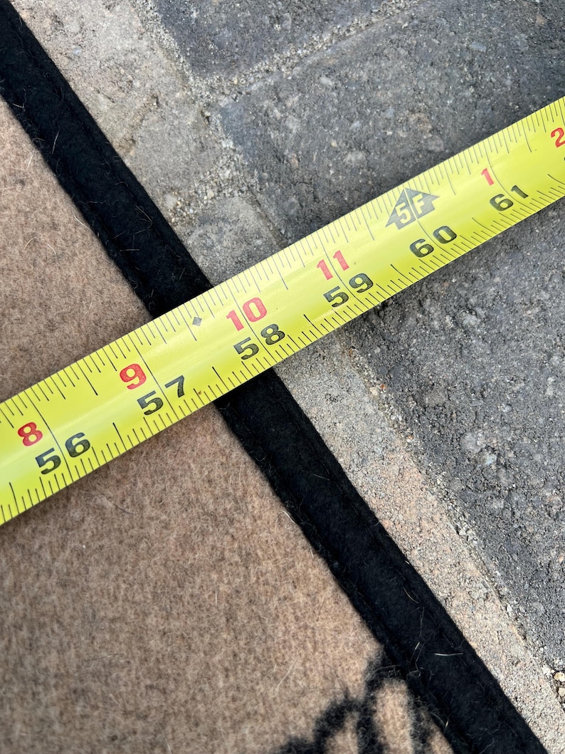 Op de afbeelding: Een beige textiel met een zwarte rand ligt onder een geel meetlint. Het lint toont markeringen van 142 tot 155 cm, wat de lengte van de rand van het beige materiaal aangeeft terwijl het uitgelijnd is met het meetinstrument in de afbeelding.
