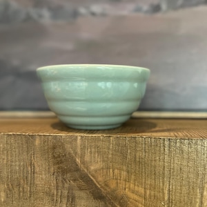 Peut inclure: Un bol en céramique vert clair avec un extérieur nervuré. Le bol est posé sur une surface en bois. L'arrière-plan est de couleur neutre et flou.