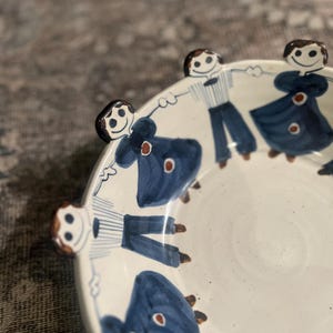 Könnte beinhalten: Eine weiße Keramikschale mit einem skurrilen Design. Der Rand zeigt kleine, dreidimensionale Figuren, die sich an den Händen halten. Das Innere der Schale ist mit blauen Figuren und roten Akzenten verziert. Das Gesamtdesign ist verspielt und künstlerisch.