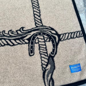 Op de afbeelding: Een beige wollen deken met een zwart grafisch patroon van verstrengelde touwen en een hoefijzer. De deken heeft een zwarte gestikte rand en een klein blauw rechthoekig label in de hoek met de witte tekst Pendleton Portland Oregon.