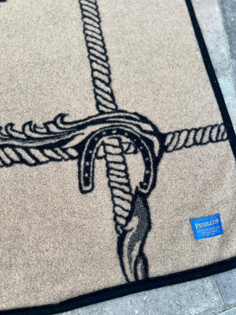 Op de afbeelding: Een beige wollen deken met een zwart grafisch patroon van verstrengelde touwen en een hoefijzer. De deken heeft een zwarte gestikte rand en een klein blauw rechthoekig label in de hoek met de witte tekst Pendleton Portland Oregon.