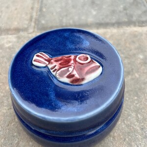 Puede incluir: Un tarro de cerámica redondo de color azul profundo con un acabado brillante. La tapa presenta un adorno de pez de cerámica tridimensional en relieve en tonos rojo oscuro y blanco, colocado en el centro de la superficie superior de la tapa.
