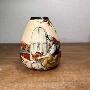 May include: A small, egg-shaped ceramic vase with a hand-painted scene. The artwork depicts a person near a well, with a teapot. The vase has a cream-colored background with brown and blue accents. The top rim is black.