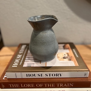 May include: A gray ceramic vase with a flared rim and a textured surface, resting on a stack of books. The books include titles like "House Story" and "The Lore of the Train". The vase has small, raised dots.