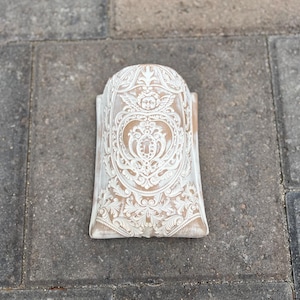May include: A semi-cylindrical wall tile featuring a light brown base with intricate white relief carvings. The design includes a central cherub figure surrounded by ornate scrollwork, floral motifs, and a keyhole shape, creating a textured surface.