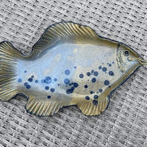 Puede incluir: Un plato de cerámica con forma de pez y esmalte azul y dorado. El plato tiene una superficie texturizada y está decorado con puntos azul oscuro. Las aletas y la cola están detalladas con un patrón acanalado. Una pieza decorativa única.
