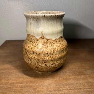 May include: A handmade ceramic vase with a speckled brown base and a creamy white top. The vase has a unique, textured surface and a slightly flared rim. The pottery is likely stoneware and is suitable for holding flowers or as a decorative piece.