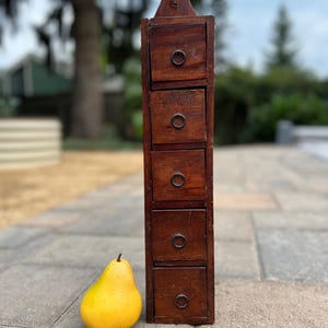 May include: A tall, narrow, dark brown wooden storage unit featuring five stacked drawers, each equipped with a small, circular metal pull handle. A single yellow pear is positioned directly to the left of the wooden cabinet on the ground.