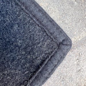 Op de afbeelding: Een hoek van donker antracietkleurig vilttextiel met een dichte, vezelige textuur waarin lichte vezels zichtbaar zijn verwerkt. De rand is afgewerkt met een dikke, gestikte boord die een verhoogde, gedefinieerde naad langs de omtrek vormt.