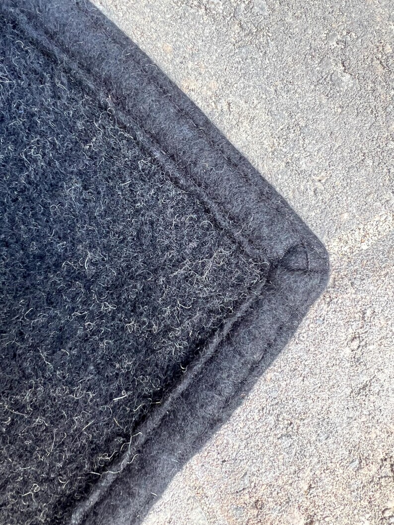 Op de afbeelding: Een hoek van donker antracietkleurig vilttextiel met een dichte, vezelige textuur waarin lichte vezels zichtbaar zijn verwerkt. De rand is afgewerkt met een dikke, gestikte boord die een verhoogde, gedefinieerde naad langs de omtrek vormt.