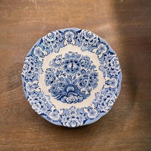 May include: A round, blue and white ceramic plate with a floral design. The plate features a central bouquet of blue flowers surrounded by a border of intricate blue patterns. The rim has a scalloped edge. The plate is set against a wooden surface.