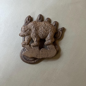 May include: A brown, textured cast metal wall plaque shaped like a bear paw print. A three-dimensional bear figure is centred on the paw, standing on a raised rock-like base. The entire piece features a weathered, rustic finish with dark brown highlights.