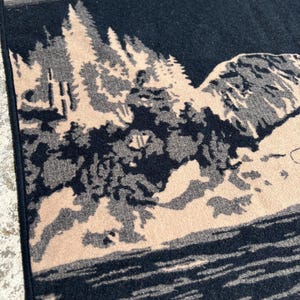 Puede incluir: Una manta textil tejida con un dise&ntilde;o de paisaje bicolor en negro y beige. El patr&oacute;n representa un denso bosque de pinos, una ladera monta&ntilde;osa rocosa y un cuerpo de agua. Una parte de una ilustraci&oacute;n de un pez es visible en el borde superior.