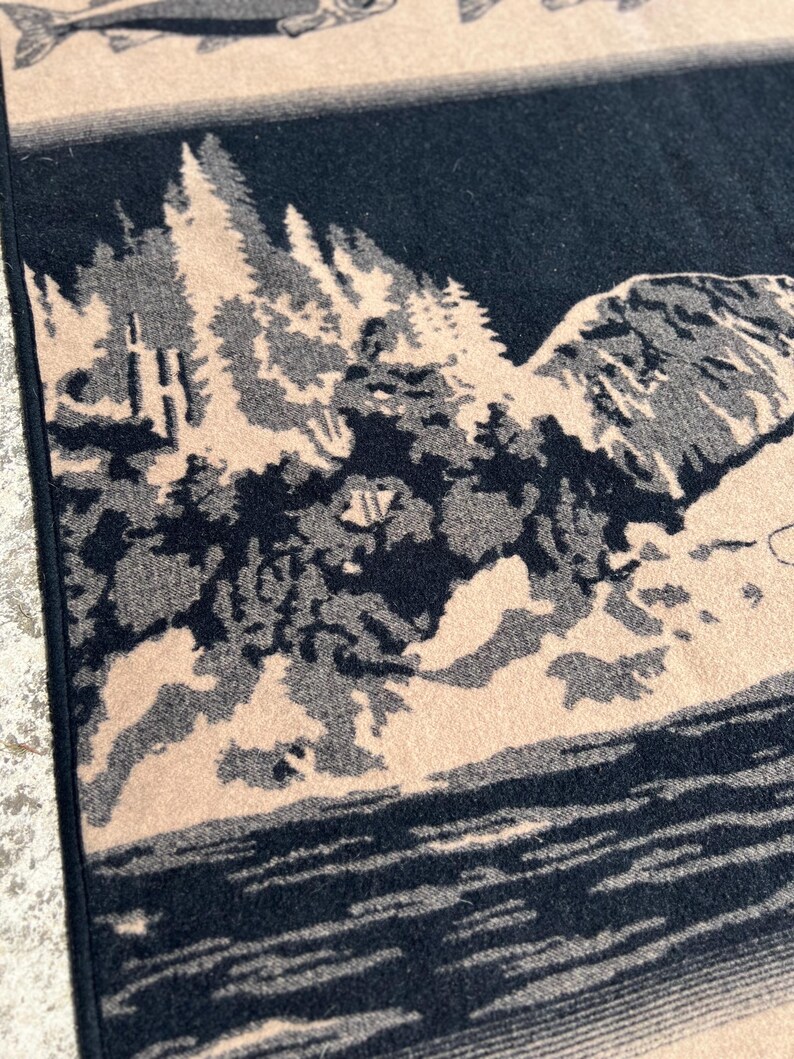 Puede incluir: Una manta textil tejida con un dise&ntilde;o de paisaje bicolor en negro y beige. El patr&oacute;n representa un denso bosque de pinos, una ladera monta&ntilde;osa rocosa y un cuerpo de agua. Una parte de una ilustraci&oacute;n de un pez es visible en el borde superior.