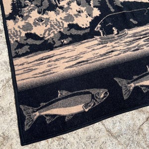 Puede incluir: Una manta tejida con una escena al aire libre en negro y beige que muestra a una persona en un bote peque&ntilde;o en un lago rodeado de &aacute;rboles y monta&ntilde;as. La secci&oacute;n inferior presenta un patr&oacute;n de peces nadando sobre un fondo oscuro.