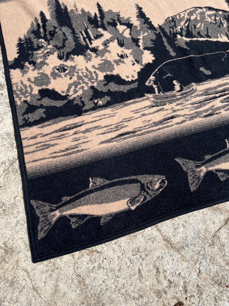 Puede incluir: Una manta tejida con una escena al aire libre en negro y beige que muestra a una persona en un bote peque&ntilde;o en un lago rodeado de &aacute;rboles y monta&ntilde;as. La secci&oacute;n inferior presenta un patr&oacute;n de peces nadando sobre un fondo oscuro.
