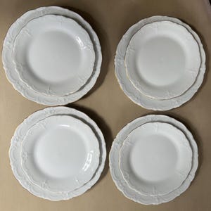 Könnte beinhalten: Vier Sets weißer Porzellanteller. Jedes Set enthält einen großen Teller und einen kleineren Teller, die übereinander gestapelt sind. Die Teller haben einen gewellten Rand und ein zartes Blumendesign. Die Teller sind auf einer neutralfarbenen Oberfläche angeordnet.