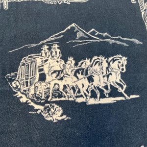 Op de afbeelding: Een donkerblauwe textiel met een witte geweven afbeelding van een postkoets getrokken door vier paarden met twee figuren bovenop. De sc&egrave;ne is geplaatst tegen een achtergrond van een bergketen en omlijst door een decoratieve touwrand.