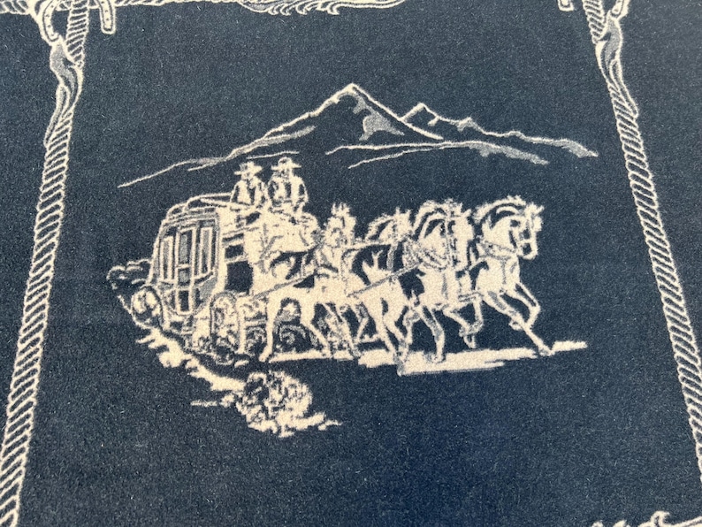 Op de afbeelding: Een donkerblauwe textiel met een witte geweven afbeelding van een postkoets getrokken door vier paarden met twee figuren bovenop. De sc&egrave;ne is geplaatst tegen een achtergrond van een bergketen en omlijst door een decoratieve touwrand.