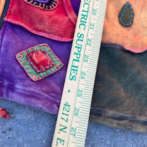 K&ouml;nnte beinhalten: Ein mehrfarbiger Patchwork-Kapuzenpullover mit roten, violetten und orangefarbenen Stoffbahnen, Ziern&auml;hten und Applikationen. Ein Holzlineal liegt vertikal auf dem Kleidungsst&uuml;ck und zeigt Ma&szlig;e von 45 bis 71 cm an.