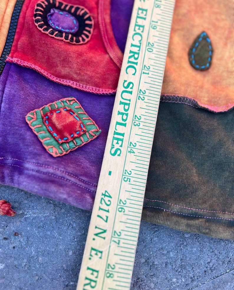 K&ouml;nnte beinhalten: Ein mehrfarbiger Patchwork-Kapuzenpullover mit roten, violetten und orangefarbenen Stoffbahnen, Ziern&auml;hten und Applikationen. Ein Holzlineal liegt vertikal auf dem Kleidungsst&uuml;ck und zeigt Ma&szlig;e von 45 bis 71 cm an.