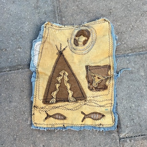May include: A rectangular fabric patch featuring a yellow base layer with dark brown felt appliqués including a teepee, a bull skull, two fish, and a circular sun motif. The edges are finished with dark zigzag stitching on a blue denim backing.