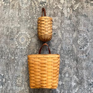 May include: Two light brown woven baskets with smooth, flat wooden splints and brown leather straps. The top basket is cylindrical and smaller, while the bottom basket is larger, rectangular, and features a wider, open-top design.