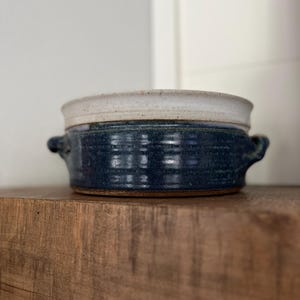 Puede incluir: Un cuenco de cerámica hecho a mano con exterior azul oscuro y borde color crema. El cuenco tiene dos asas pequeñas y está sobre una superficie de madera. El cuenco de gres es adecuado para servir sopas, guisos u otros platos.