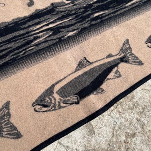 Puede incluir: Un tejido de color tostado con un estampado gr&aacute;fico negro de un pez nadando en primer plano y un barco con figuras sobre el agua al fondo. La tela tiene un borde negro oscuro a lo largo del margen, mostrando un patr&oacute;n tejido detallado.