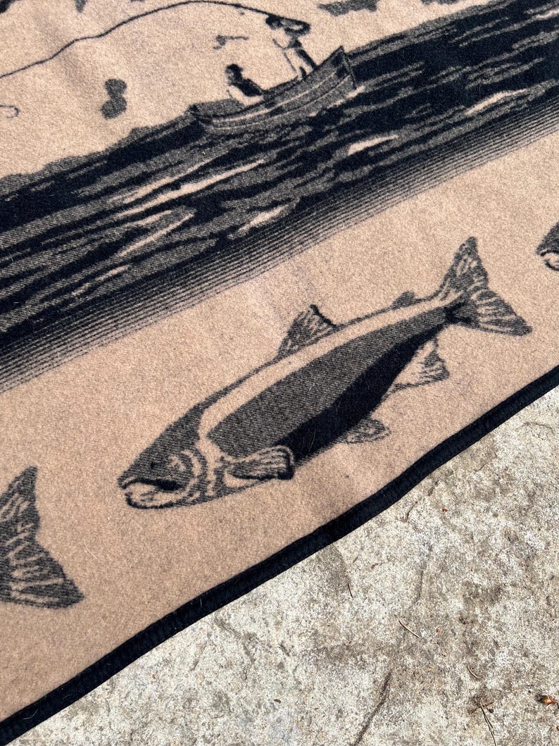 Puede incluir: Un tejido de color tostado con un estampado gr&aacute;fico negro de un pez nadando en primer plano y un barco con figuras sobre el agua al fondo. La tela tiene un borde negro oscuro a lo largo del margen, mostrando un patr&oacute;n tejido detallado.