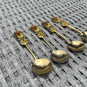 Puede incluir: Un juego de cuatro cucharas decorativas con cuencos y mangos dorados. Los mangos de las cucharas tienen una parte superior rectangular, negra y decorativa con un diseño floral colorido. Las cucharas están dispuestas sobre una superficie tejida gris.