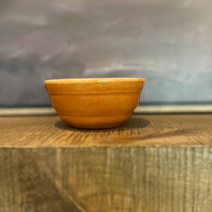 Può includere: Una piccola ciotola rotonda in ceramica arancione con un bordo leggermente svasato. La ciotola ha una finitura liscia e lucida ed è appoggiata su una superficie di legno. La ciotola viene probabilmente utilizzata per servire o mescolare ingredienti.