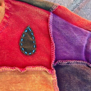 K&ouml;nnte beinhalten: Ein Patchwork-Sweatshirt mit Stoffbahnen in Rot, Orange, Violett und Braun, die durch sichtbare rosa Overlockn&auml;hte verbunden sind. Ein dunkelbrauner, tropfenf&ouml;rmiger Aufn&auml;her ist mit hellblauen Handstickereien auf dem roten Teil befestigt.