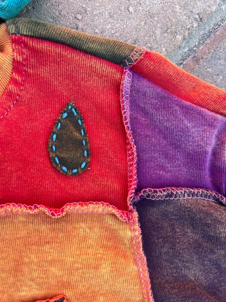 K&ouml;nnte beinhalten: Ein Patchwork-Sweatshirt mit Stoffbahnen in Rot, Orange, Violett und Braun, die durch sichtbare rosa Overlockn&auml;hte verbunden sind. Ein dunkelbrauner, tropfenf&ouml;rmiger Aufn&auml;her ist mit hellblauen Handstickereien auf dem roten Teil befestigt.