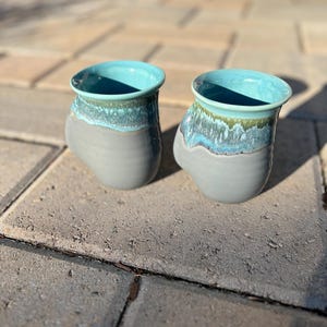 May include: Two handmade ceramic mugs. The mugs have a gray base with a turquoise interior and a turquoise and green rim. The mugs are sitting on a gray brick surface.