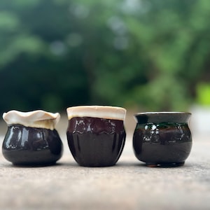 May include: Three small, handcrafted ceramic pots in a row. The pots are dark brown with unique details: one has a ruffled white rim, another has a tan rim, and the third has a green rim. The pots are likely for small plants or succulents.