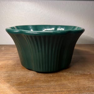 May include: A dark green ceramic bowl with a fluted exterior. The bowl has a slightly flared rim and sits on a wooden surface. The bowl is empty and appears to be unused. The bowl is a decorative item.
