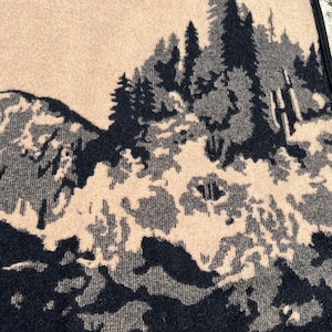 Puede incluir: Una manta tejida con un patr&oacute;n de paisaje en alto contraste negro y beige. El dise&ntilde;o muestra un bosque denso de pinos frente a picos monta&ntilde;osos escarpados, con un borde negro s&oacute;lido en la parte superior y un acabado de tela texturizada.